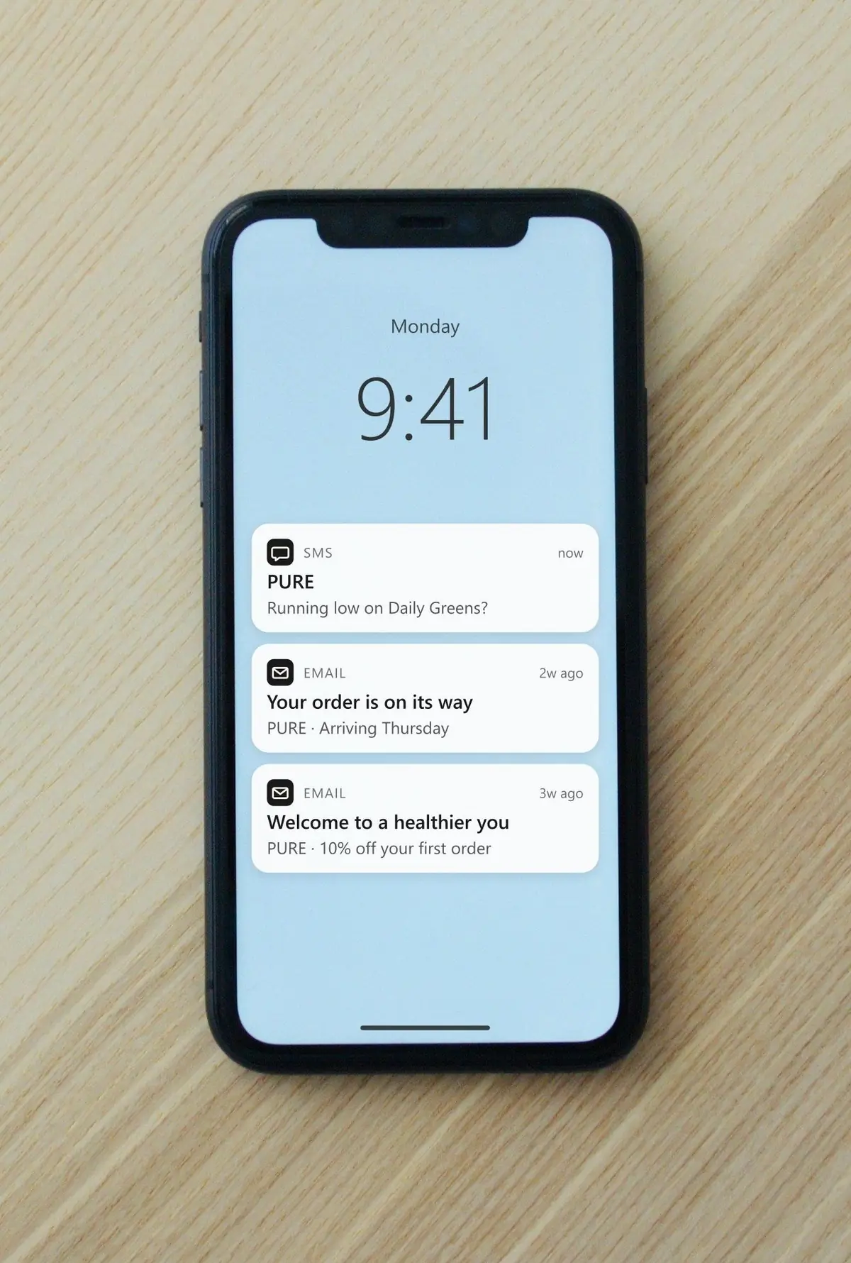 A phone on a wooden desk with three notifications from an online store: a restock reminder by text, a shipping update and a welcome offer by email
